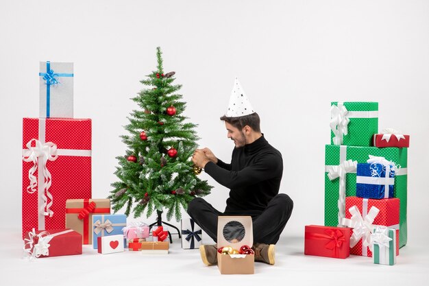 Vista frontal del joven sentado alrededor de regalos de vacaciones decorando arbolito en la pared blanca