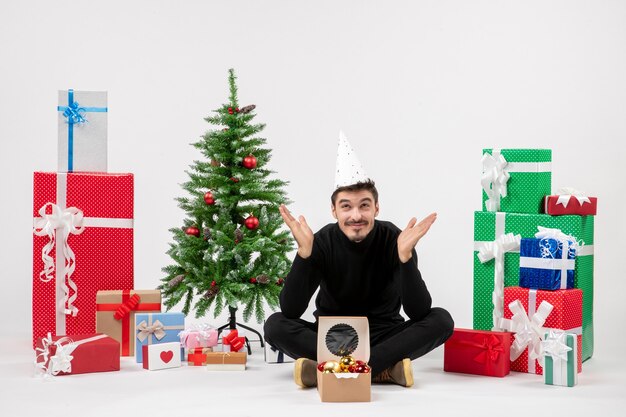 Vista frontal del joven sentado alrededor de regalos de vacaciones en la pared blanca