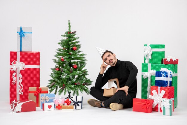 Vista frontal del joven sentado alrededor de regalos de vacaciones en la pared blanca