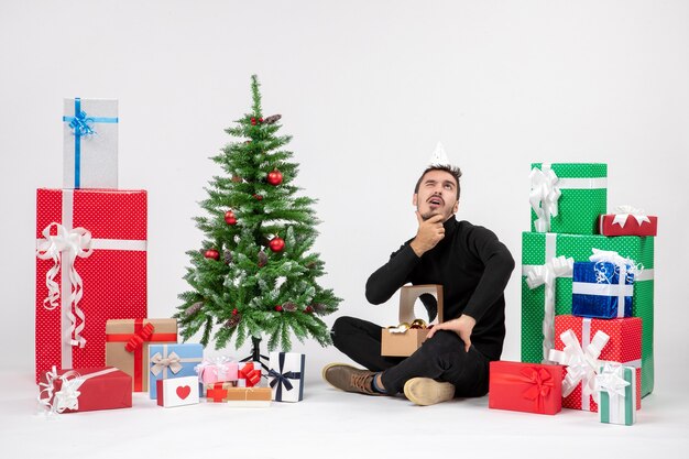 Vista frontal del joven sentado alrededor de regalos de vacaciones en la pared blanca