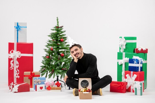 Vista frontal del joven sentado alrededor de regalos de vacaciones en la pared blanca