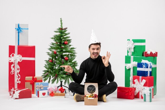 Vista frontal del joven sentado alrededor de regalos de vacaciones en la pared blanca