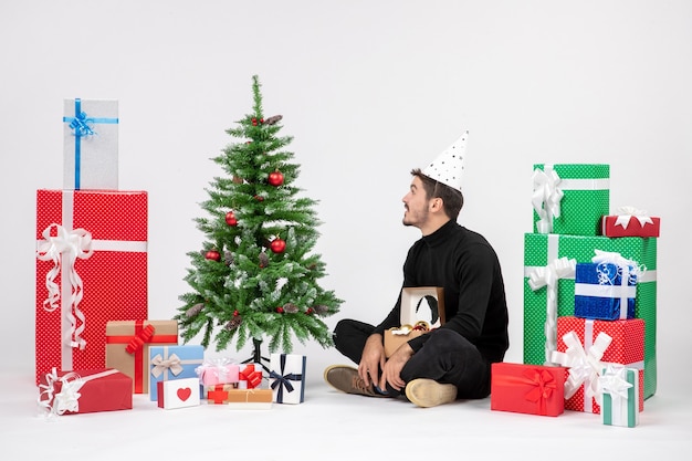 Vista frontal del joven sentado alrededor de regalos de vacaciones en la pared blanca