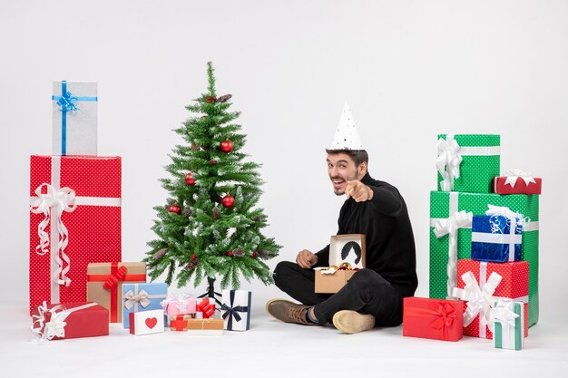 Vista frontal del joven sentado alrededor de regalos de vacaciones en la pared blanca