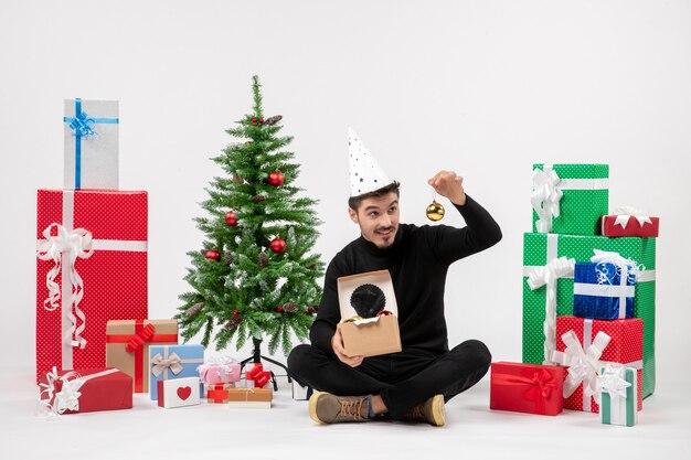 Vista frontal del joven sentado alrededor de regalos de vacaciones en la pared blanca