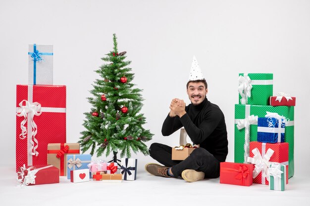 Vista frontal del joven sentado alrededor de regalos de vacaciones en la pared blanca