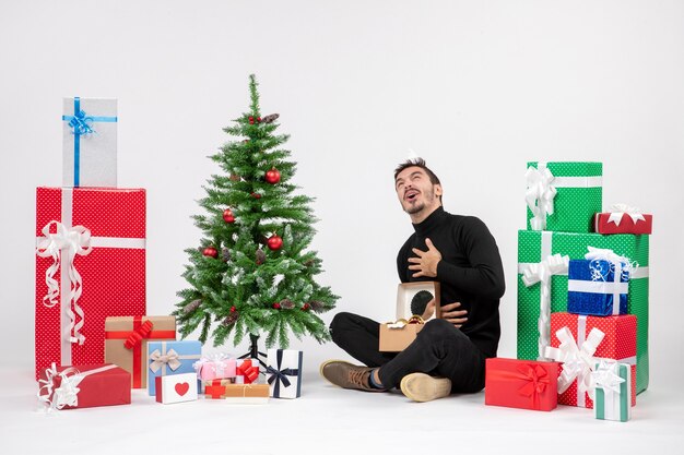 Vista frontal del joven sentado alrededor de regalos de vacaciones en la pared blanca