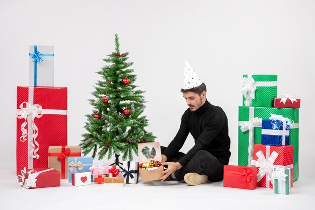 Vista frontal del joven sentado alrededor de regalos de vacaciones sosteniendo el paquete con juguetes en la pared blanca