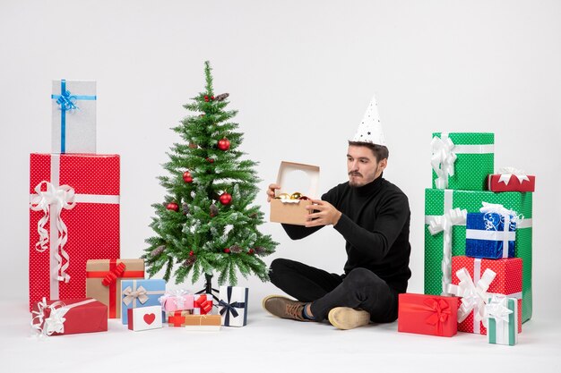 Vista frontal del joven sentado alrededor de regalos de vacaciones sosteniendo el paquete con juguetes en la pared blanca