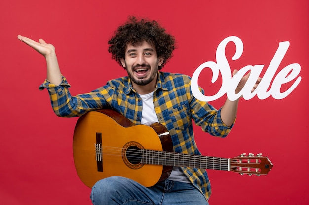 Vista frontal joven sentado con guitarra en pared roja concierto aplauso músico en vivo venta de colores