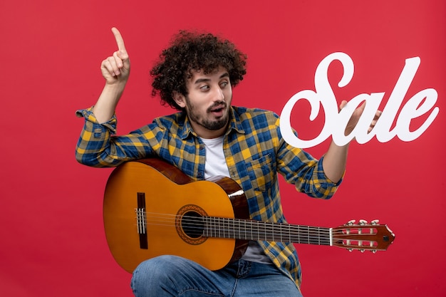 Foto gratuita vista frontal joven sentado con la guitarra en la pared roja concierto de música aplausos músico en vivo venta de colores