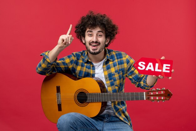 Vista frontal joven sentado con guitarra en pared roja tocar música músico concierto en vivo de color