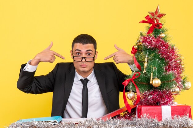 Vista frontal joven sentado en la mesa apuntando con el dedo a sí mismo árbol de Navidad y regalos