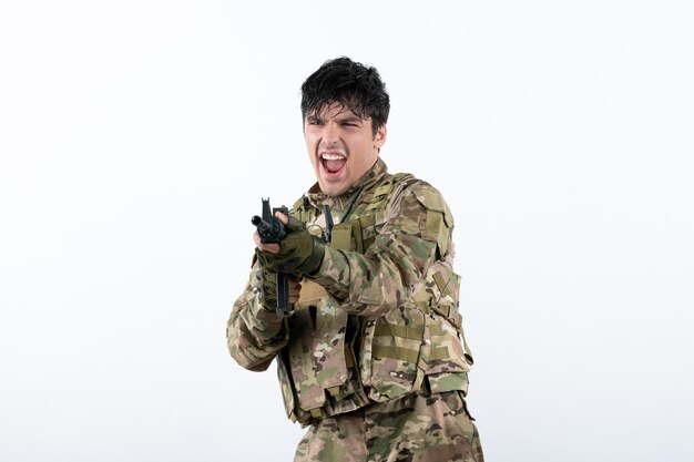 Vista frontal del joven soldado en camuflaje con ametralladora pared blanca