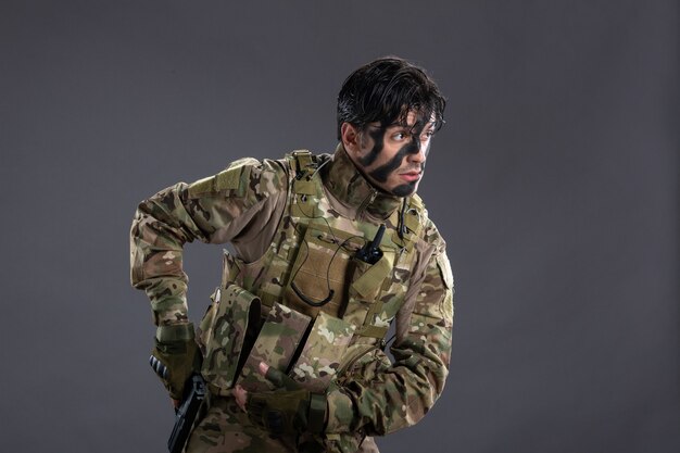 Vista frontal del joven soldado de camuflaje con pistola en la pared oscura