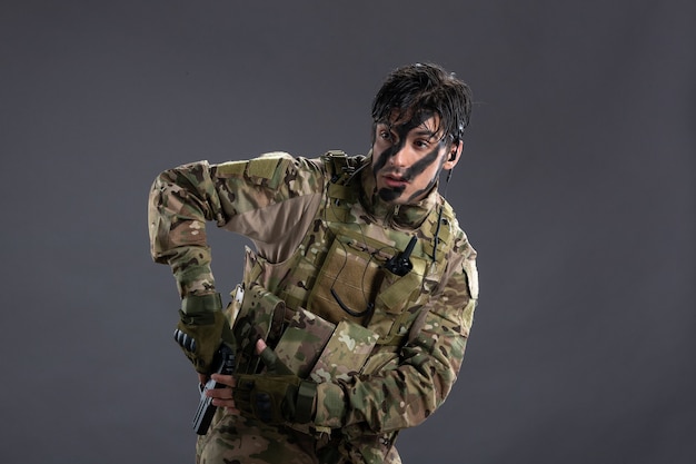 Vista frontal del joven soldado de camuflaje con pistola en la pared oscura