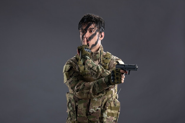 Vista frontal del joven soldado de camuflaje con pistola en la pared oscura