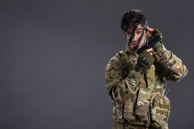 Vista frontal del joven soldado de camuflaje con pistola en la pared oscura