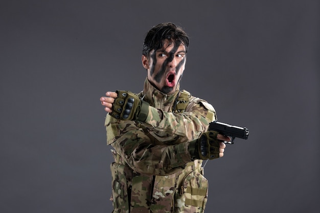 Vista frontal del joven soldado de camuflaje con pistola en la pared oscura