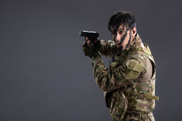 Vista frontal joven soldado luchando en camuflaje con pistola en la pared oscura