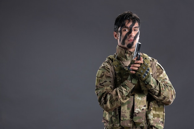 Vista frontal joven soldado luchando en camuflaje con pistola en la pared oscura