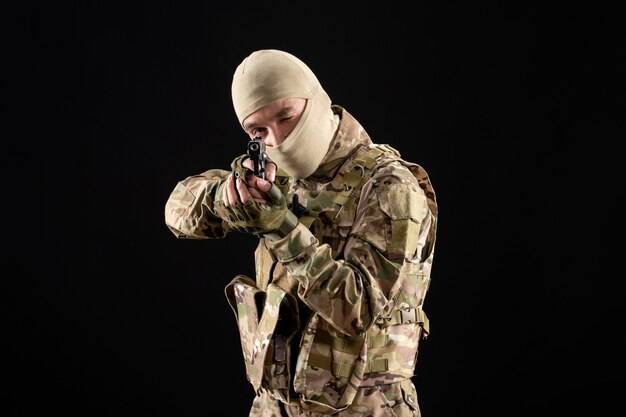 Vista frontal del joven soldado en uniforme apuntando con pistola en la pared negra