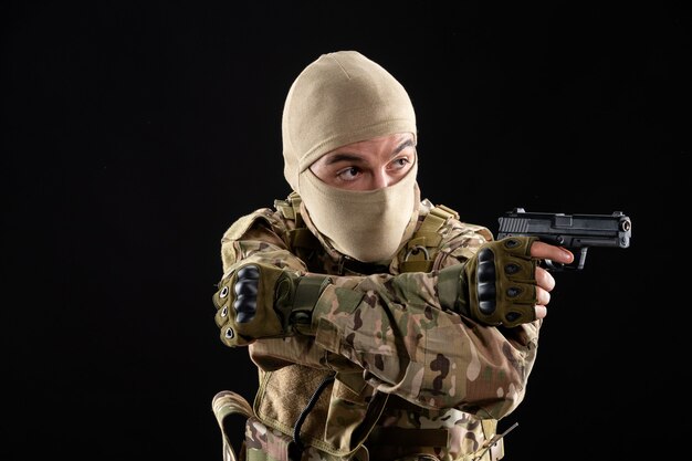 Vista frontal del joven soldado en uniforme apuntando con pistola en la pared negra