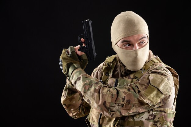 Vista frontal del joven soldado en uniforme apuntando con pistola en la pared negra