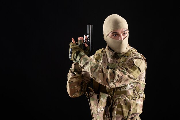 Vista frontal del joven soldado en uniforme apuntando con pistola en la pared negra
