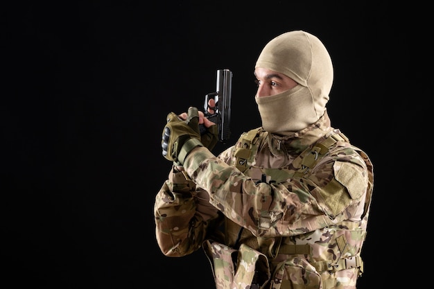 Vista frontal del joven soldado en uniforme apuntando con pistola en la pared negra