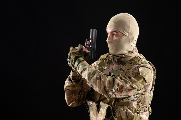 Vista frontal del joven soldado en uniforme apuntando con pistola en la pared negra