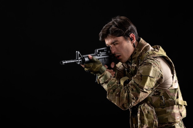 Vista frontal joven soldado en uniforme apuntando con su rifle en la pared negra