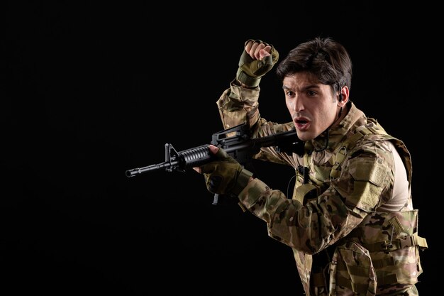 Vista frontal joven soldado en uniforme apuntando con su rifle en la pared negra