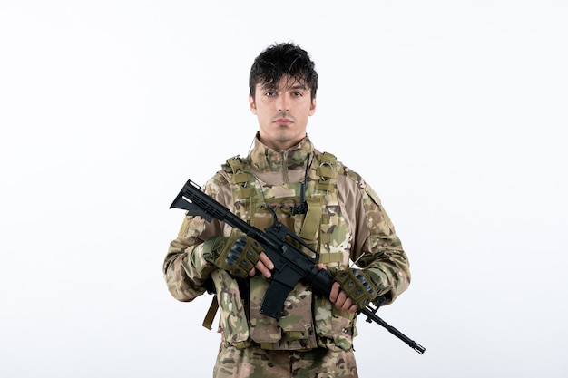 Vista frontal del joven soldado en uniforme militar con ametralladora pared blanca