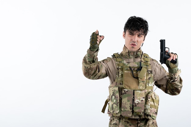 Vista frontal joven soldado en uniforme militar con pistola pared blanca