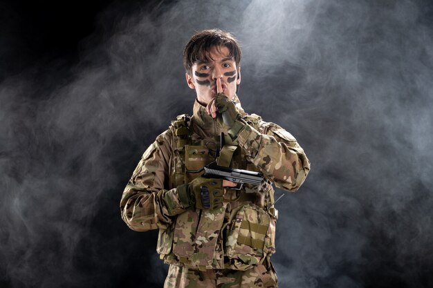 Vista frontal del joven soldado en uniforme con pistola en la pared negra