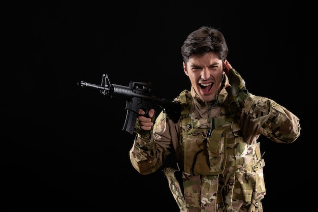 Vista frontal joven soldado en uniforme con rifle en la pared negra