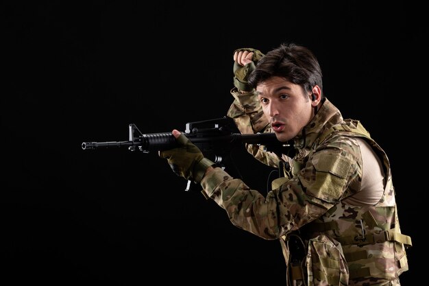 Vista frontal joven soldado en uniforme con rifle en la pared negra