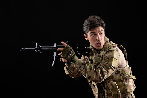 Vista frontal joven soldado en uniforme con rifle en la pared negra