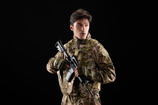 Vista frontal del joven soldado en uniforme con rifle en pared negra