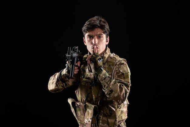 Vista frontal del joven soldado en uniforme con rifle en pared negra