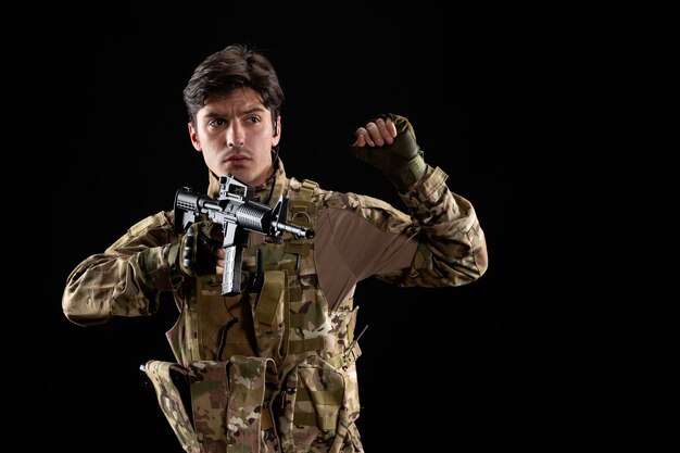 Vista frontal del joven soldado en uniforme con rifle en pared negra