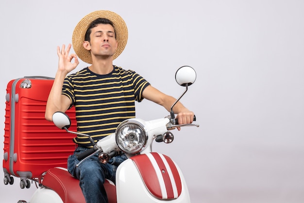 Vista frontal del joven con sombrero de paja en ciclomotor gesticulando ok firmar cerrando los ojos