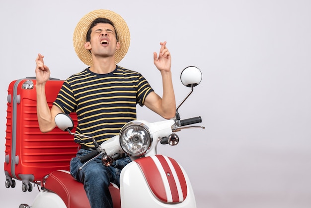 Vista frontal del joven con sombrero de paja en ciclomotor haciendo deseo firmar con los ojos cerrados