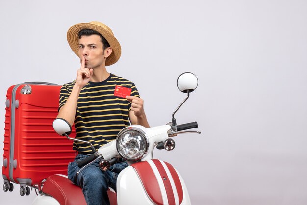 Vista frontal del joven con sombrero de paja en ciclomotor con tarjeta de crédito haciendo signo shh