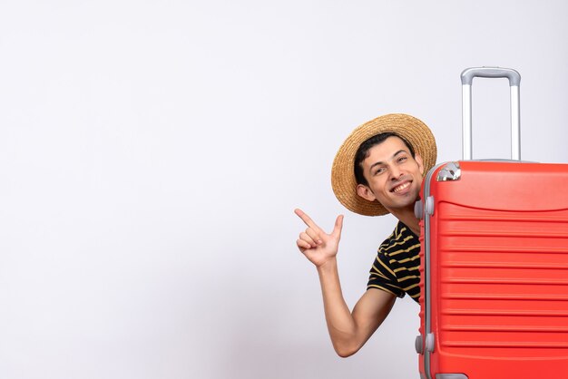 Vista frontal joven con sombrero de paja de pie detrás de la maleta roja apuntando a algo