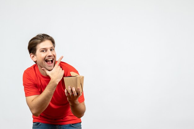 Vista frontal del joven sonriente en blusa roja sosteniendo una pequeña caja sobre fondo blanco.