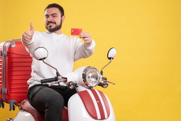 Vista frontal del joven sonriente hombre que viaja sentado en la motocicleta con la maleta en ella sosteniendo una tarjeta bancaria haciendo un gesto bien sobre fondo amarillo aislado