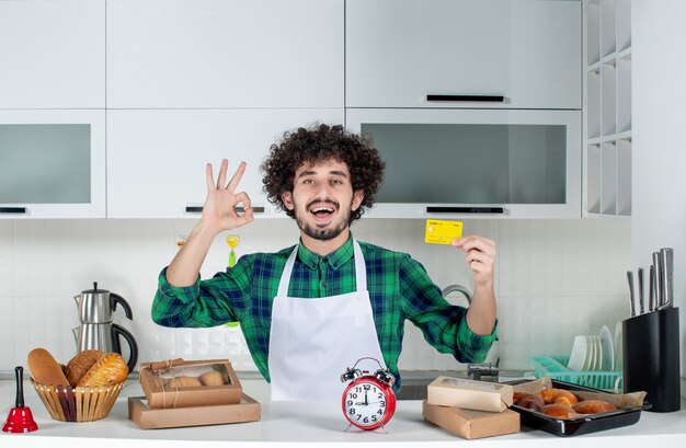 Vista frontal del joven sonriente de pie detrás de la mesa con varios pasteles y sosteniendo una tarjeta bancaria y haciendo un gesto de ok en la cocina blanca