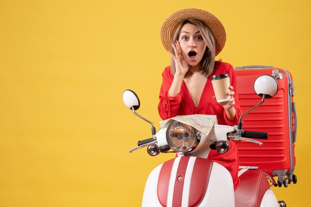 Vista frontal de la joven sorprendida en vestido rojo sosteniendo la taza de café cerca del ciclomotor